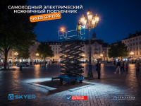 Ножничный подъемник SKYER SPF1305 ночью на городской площади с включенным рабочим освещением. Оператор на платформе выполняет ремонт уличного освещения.
